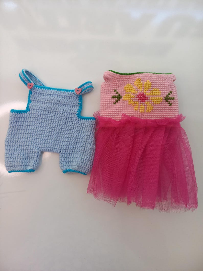 May include: Two crocheted doll outfits. One is a light blue overall with blue trim and pink heart-shaped buttons. The other is a pink dress with a yellow flower design and a pink tulle skirt. The outfits are handmade.