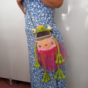 May include: A green and pink crochet frog-shaped crossbody bag with a yellow crown. The bag has a pink tutu skirt and long green legs. The bag is hanging from a silver chain. The bag is holding a cell phone.
