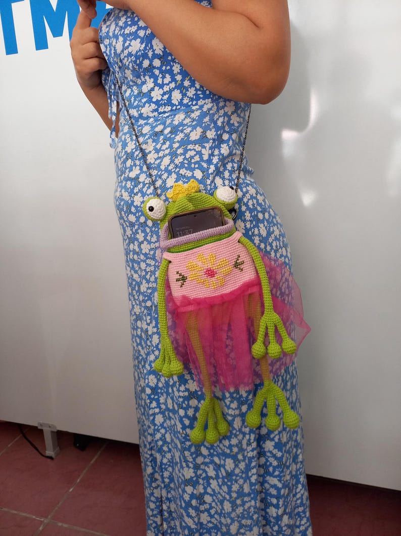 May include: A green and pink crochet frog-shaped crossbody bag with a yellow crown. The bag has a pink tutu skirt and long green legs. The bag is hanging from a silver chain. The bag is holding a cell phone.