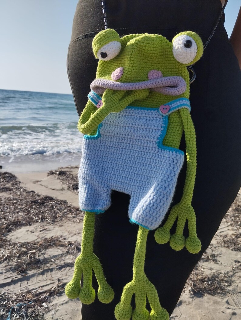 Peut inclure: Sac en crochet fait main en forme de grenouille. La t&ecirc;te de la grenouille est verte avec de grands yeux blancs, des joues roses et une bouche lavande. Elle porte une salopette bleu clair avec des bordures turquoise et des boutons roses en forme de c&oelig;ur. Les longues pattes vertes de la grenouille pendent.