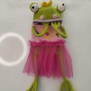 May include: A handmade crochet frog doll wearing a pink tutu and a yellow crown. The frog is primarily green with white and black eyes, and pink accents. The doll's body is pink with yellow cross-stitch flowers.