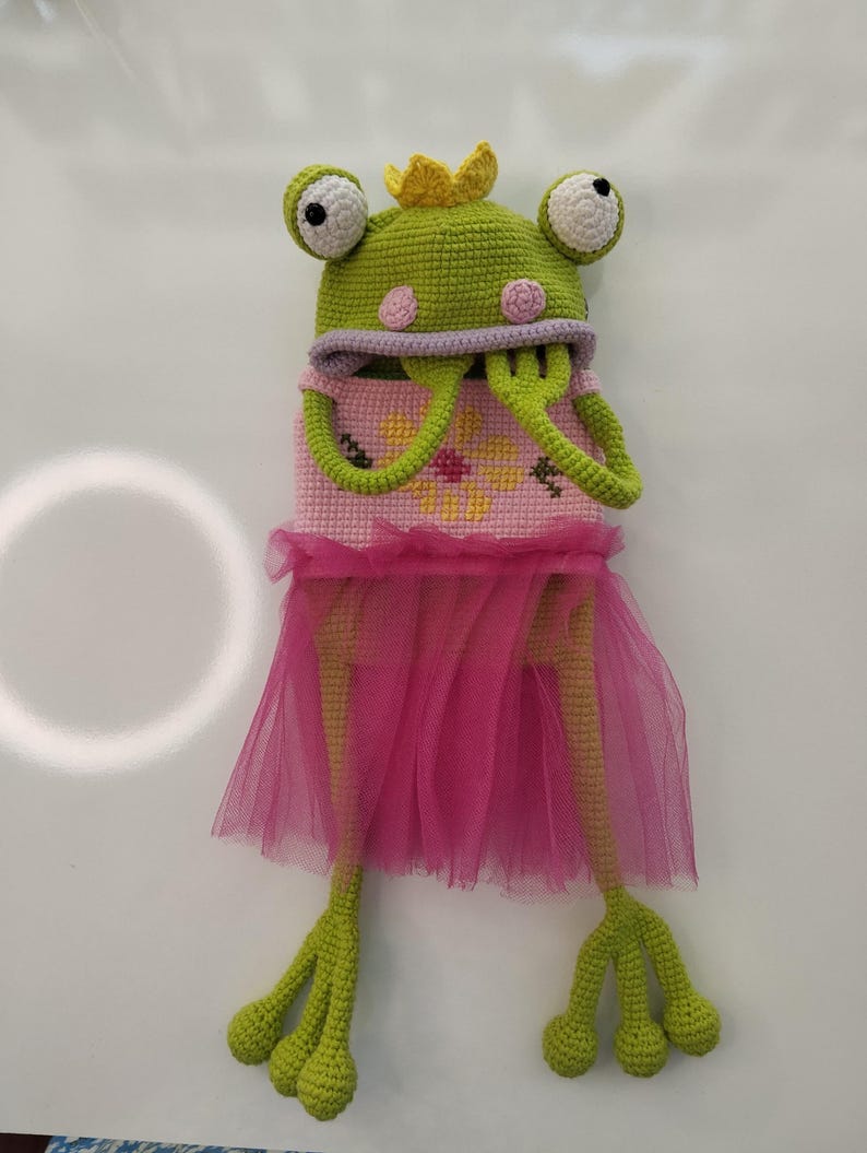 May include: A handmade crochet frog doll wearing a pink tutu and a yellow crown. The frog is primarily green with white and black eyes, and pink accents. The doll's body is pink with yellow cross-stitch flowers.