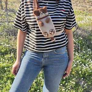 May include: A beige and brown crochet cat-shaped crossbody bag. The bag has a long strap and features a cat face, paws, and a tail. The person is wearing a black and white striped t-shirt and blue jeans. The word "Happy" is embroidered on the shirt.