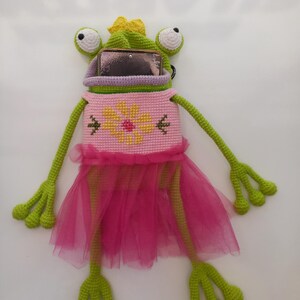 May include: A whimsical, handmade frog-shaped phone case. The case is primarily green, with a pink dress and a yellow flower design on the front. The frog has large, white and black eyes and a yellow bow.