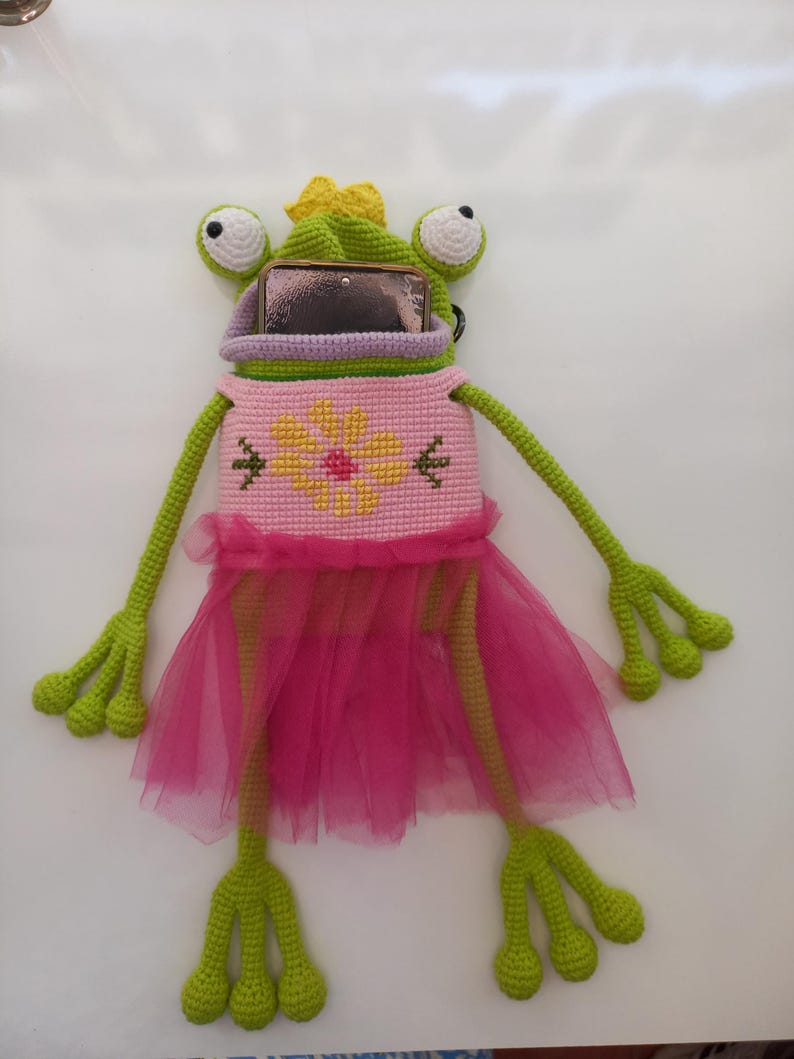May include: A whimsical, handmade frog-shaped phone case. The case is primarily green, with a pink dress and a yellow flower design on the front. The frog has large, white and black eyes and a yellow bow.
