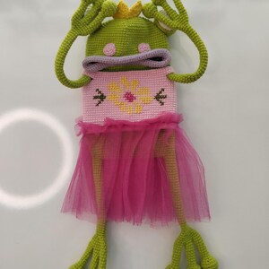 May include: A whimsical, handmade crochet frog doll. The frog is green with large eyes, a crown, and long arms. It wears a pink dress with a flower design and a tulle skirt. The doll is a unique, handcrafted item.
