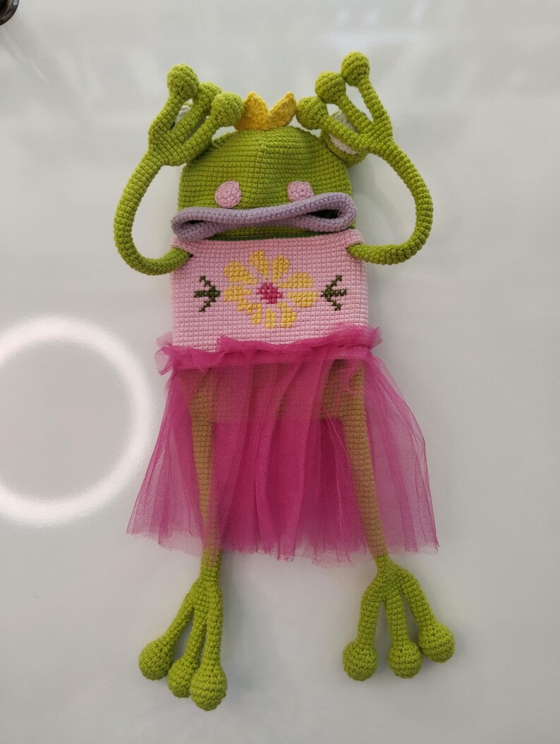 May include: A whimsical, handmade crochet frog doll. The frog is green with large eyes, a crown, and long arms. It wears a pink dress with a flower design and a tulle skirt. The doll is a unique, handcrafted item.