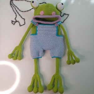 Peut inclure: Une poup&eacute;e grenouille au crochet faite &agrave; la main, avec une t&ecirc;te verte, des joues roses et de grands yeux bord&eacute;s de blanc. Elle porte une salopette bleu clair avec des bordures bleues et a de longs bras et jambes verts. Une cha&icirc;ne d&eacute;corative est &agrave; proximit&eacute;.