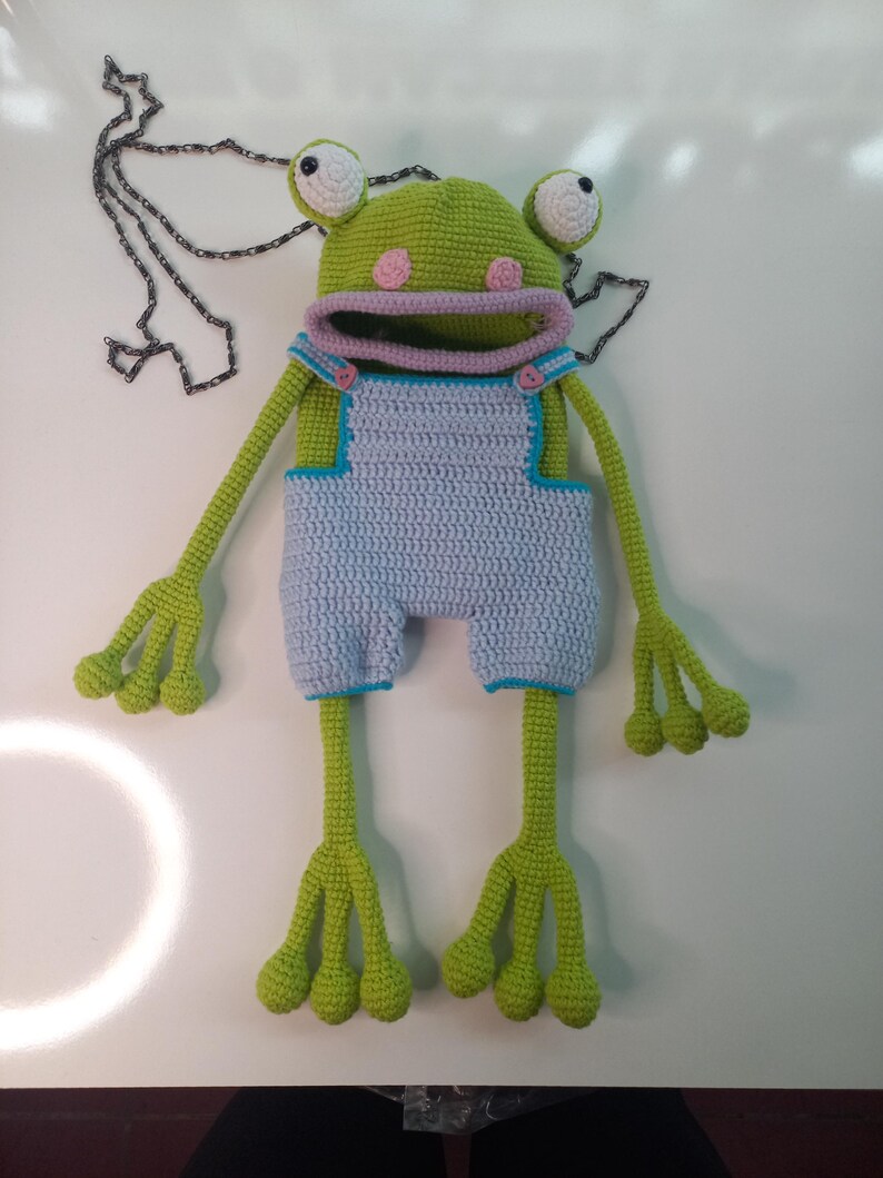 Peut inclure: Une poup&eacute;e grenouille au crochet faite &agrave; la main, avec une t&ecirc;te verte, des joues roses et de grands yeux bord&eacute;s de blanc. Elle porte une salopette bleu clair avec des bordures bleues et a de longs bras et jambes verts. Une cha&icirc;ne d&eacute;corative est &agrave; proximit&eacute;.