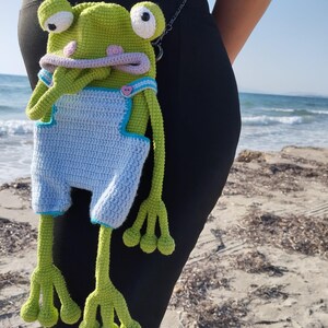 Peut inclure: Un sac en forme de grenouille au crochet fait &agrave; la main, dans des tons de vert, de bleu et de lavande. Le sac en forme de grenouille pr&eacute;sente de grands yeux, une large bouche et une salopette bleue. Le sac a de longues pattes et une bandouli&egrave;re en cha&icirc;ne.