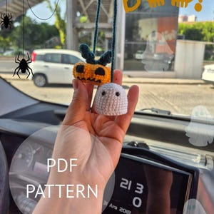 May include: Crocheted Halloween car charm featuring a pumpkin and a ghost. The pumpkin is orange with green leaves, and the ghost is white. The charm hangs from a green string. The word "Boo" is written in orange.