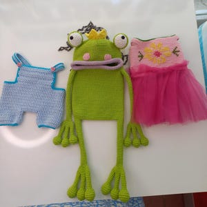 May include: A collection of handmade crochet items, including a light blue overall, a green frog hat with a yellow crown, and a pink dress with a flower design and a tulle skirt. The items are displayed on a white surface.