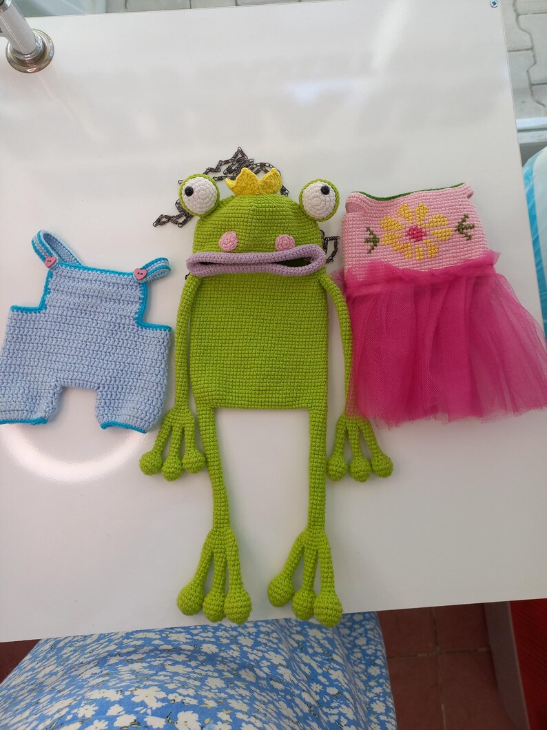 May include: A collection of handmade crochet items, including a light blue overall, a green frog hat with a yellow crown, and a pink dress with a flower design and a tulle skirt. The items are displayed on a white surface.