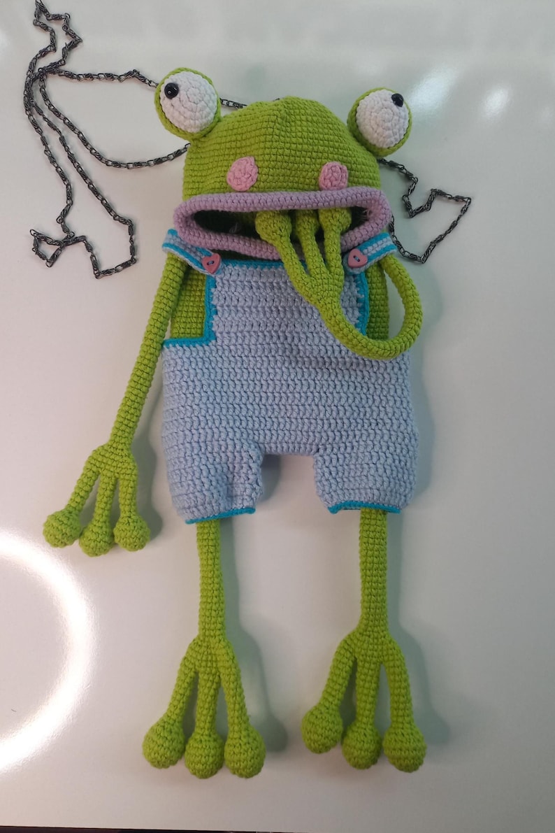 Peut inclure: Une cr&eacute;ature en forme de grenouille faite au crochet, avec de grands yeux, une t&ecirc;te verte et de longs membres. Elle porte une salopette bleu clair avec des accents roses et une cha&icirc;ne. Le design fantaisiste pr&eacute;sente une esth&eacute;tique unique et ludique.