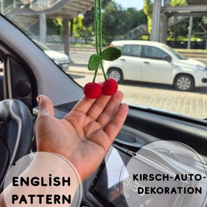 Puede incluir: Adorno de coche de ganchillo con dos cerezas rojas con hojas verdes y un tallo verde. El adorno cuelga en un coche, con el texto "English Pattern" visible. El fondo muestra una calle.