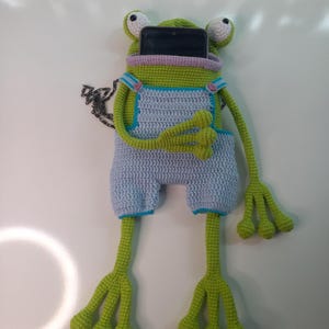 Peut inclure: Une housse de t&eacute;l&eacute;phone en crochet en forme de grenouille, faite &agrave; la main, de couleur verte, bleu clair et rose. La grenouille a de grands yeux et de longues pattes, avec une poche pour un t&eacute;l&eacute;phone.