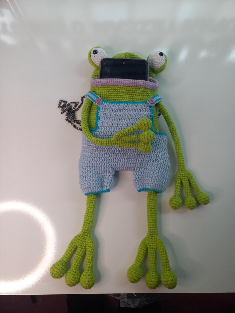 Peut inclure: Une housse de t&eacute;l&eacute;phone en crochet en forme de grenouille, faite &agrave; la main, de couleur verte, bleu clair et rose. La grenouille a de grands yeux et de longues pattes, avec une poche pour un t&eacute;l&eacute;phone.