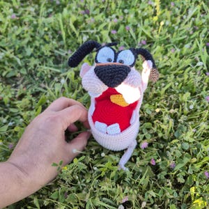 May include: A handmade crochet Courage the Cowardly Dog plushie. The plushie features Courage's signature pink, white, black, and red color scheme. The dog is in an open-mouthed expression, with a yellow tongue and white teeth. The plushie is set against a background of green grass.