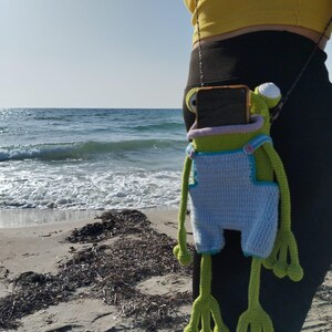 Peut inclure: &Eacute;tui de t&eacute;l&eacute;phone en forme de grenouille crochet&eacute;e, vert, bleu et blanc. L'&eacute;tui a de longues pattes et bras, et une petite cha&icirc;ne. La grenouille porte une salopette bleue. L'&eacute;tui est suspendu &agrave; une personne.