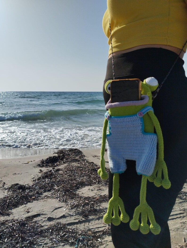 Peut inclure: &Eacute;tui de t&eacute;l&eacute;phone en forme de grenouille crochet&eacute;e, vert, bleu et blanc. L'&eacute;tui a de longues pattes et bras, et une petite cha&icirc;ne. La grenouille porte une salopette bleue. L'&eacute;tui est suspendu &agrave; une personne.