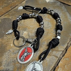 Op de afbeelding: Zwarte paracord sleutelhanger en rozenkrans met zilverkleurige kralen en Saint Michael-hangers. De ovale hangers tonen een afbeelding van Saint Michael en de woorden "Saint Michael". De sleutelhanger heeft een zilverkleurige sluiting.