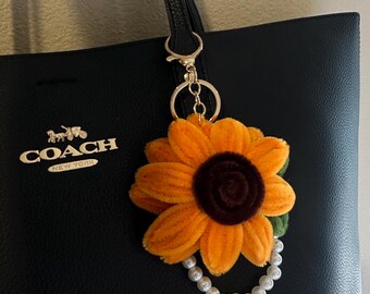 May include: A black leather tote bag with the Coach logo and a gold sunflower keychain with a pearl chain.