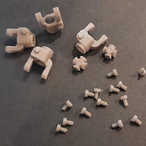May include: A selection of small, light tan 3D-printed mechanical components, including U-shaped parts, small cross-shaped pieces, and numerous tiny screws. The parts are scattered on a dark grey surface.