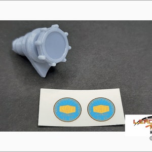 May include: A gray plastic model part with a round, textured top and a small, flat base. The part is designed to be used in model building and has a blue sticker with the word "SIATA" printed on it.