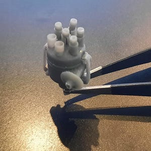 Puede incluir: Un pequeño objeto cilíndrico gris con múltiples protuberancias verticales y redondeadas. El objeto está sujeto por un par de pinzas plateadas. La sombra del objeto se proyecta sobre una superficie marrón.