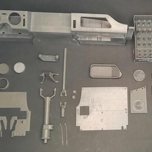 Può includere: Un assortimento di parti di modelli in plastica grigia, tra cui un cruscotto, vari pannelli, componenti circolari e piccoli pezzi rettangolari. I componenti sono disposti su una superficie scura, suggerendo un kit di modelli.