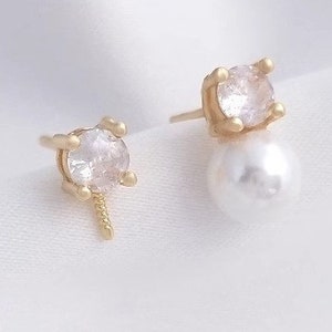 May include: A pair of gold earrings with a clear crystal and a white pearl. The earrings are on a white background.
