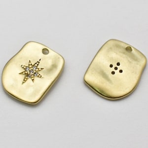 Puede incluir: Dos dijes de metal dorado, uno con un diseño en forma de estrella con diamantes de imitación transparentes y el otro con un grupo de seis agujeros.