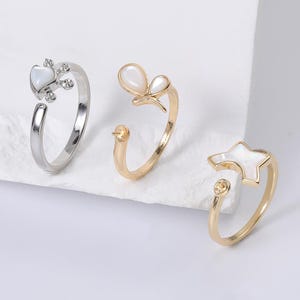 May include: Three open-ended rings with decorative elements. One ring is silver with a heart-shaped design and clear accents. Another is gold with a butterfly design. The third ring is gold with a star-shaped design and a small, round accent.