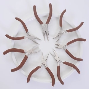 Può includere: Set di otto pinze a becchi fini con manici marroni disposte in cerchio su un piatto bianco. Le pinze sono utilizzate per la creazione di gioielli.