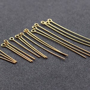 May include: A collection of gold-colored eye pins of varying lengths, arranged on a dark gray surface. The pins have a looped end and a straight wire shaft, ideal for jewelry making and crafting projects. The pins are made of metal.