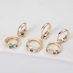 以下が含まれることがあります： カラフルな宝石が付いたゴールドトーンのフープピアス。エメラルドグリーン、サファイアブルー、ルビーレッドの石など、さまざまなデザインがあります。ピアスは明るい背景に配置されています。