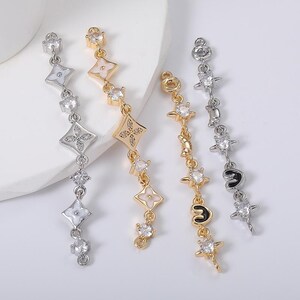 May include: Four dangling earrings in gold and silver tones. The earrings feature various designs, including diamond shapes, floral patterns, and starbursts, all adorned with clear gemstones. The earrings are displayed against a white background.
