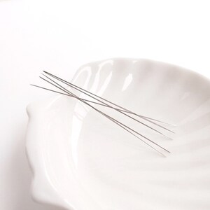 May include: Five thin, silver needles are arranged in a white ceramic bowl. The needles are straight and pointed.