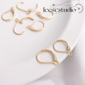May include: Gold-toned earring hooks, also known as leverback earring findings, are displayed on a white surface. The hooks are made of metal and have a smooth finish.