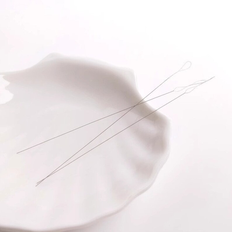 May include: Two thin, silver-colored threading needles are lying on a white ceramic dish. The needles are crossed over each other.