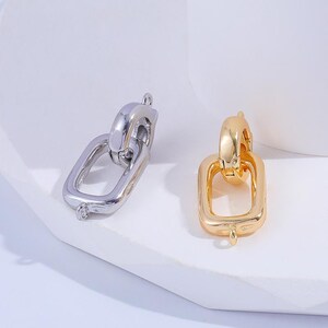 May include: Two metal jewelry connectors, one silver-toned and one gold-toned, each featuring a small circular hoop attached to a larger rectangular link. The smooth, polished surfaces reflect light, highlighting the simple, functional design of these pieces.
