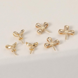 Puede incluir: Seis hallazgos de joyería dorados en forma de libélula. Las alas de la libélula están detalladas con un patrón texturizado. Cada hallazgo tiene un poste que se extiende desde el cuerpo, diseñado para sujetarse a otros componentes de joyería. Los hallazgos están sobre un fondo claro.