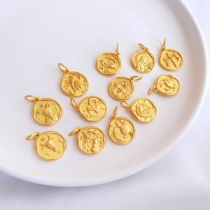 May include: Twelve gold-toned zodiac sign charms, each featuring a different astrological symbol. The charms are arranged on a white plate.