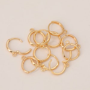May include: A group of ten gold-toned earring hoops with a hinged closure. The hoops are arranged in a pile on a white surface.