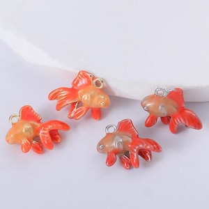 Peut inclure: Quatre breloques de poissons rouges orange et dorés, avec des boucles argentées pour la fixation. Les breloques ont des nageoires et des queues détaillées, sur fond blanc.