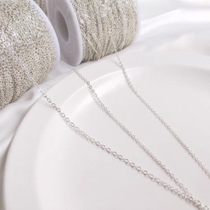 May include: Close-up of silver-colored chain on a white surface. The chain is in a continuous loop, with several spools of chain visible in the background. The chain is made of small, oval-shaped links.