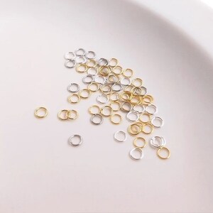 Könnte beinhalten: Nahaufnahme einer weißen Oberfläche mit einer Streuung kleiner goldener und silberner Sprungringe. Die Ringe sind alle gleich groß und in einem zufälligen Muster angeordnet.