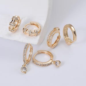 以下が含まれることがあります： ゴールドカラーのフープピアスコレクション。小さな透明な宝石で飾られたものもあれば、シンプルで洗練されたデザインのものもあります。1組にはぶら下がっている宝石が付いています。ピアスは白い表面に展示されています。
