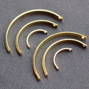 Puede incluir: Una colección de hallazgos de joyería de metal dorado y curvado. Las piezas varían en tamaño, con forma de semicírculo y pequeños agujeros en los extremos. Los hallazgos están dispuestos sobre una superficie gris oscuro, mostrando su acabado pulido.