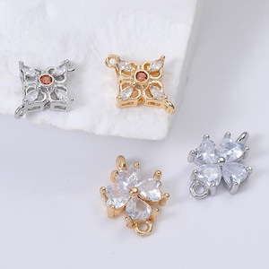 May include: Four decorative charms in gold and silver tones. Two charms have a four-petal design with clear and orange-red stones. The other two charms are clover-shaped with clear stones. Each charm has a small loop for attaching to jewelry.